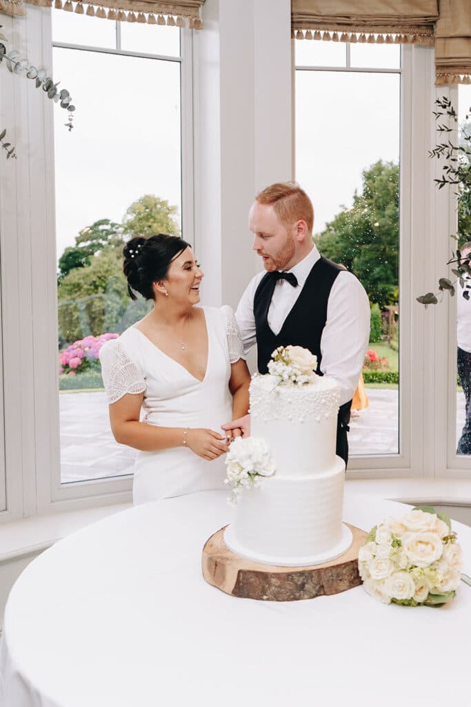 Bride and groom smiling beside white wedding cake by window