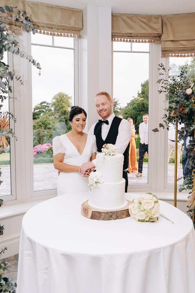 Bride and groom cutting wedding cake beside window with flowers